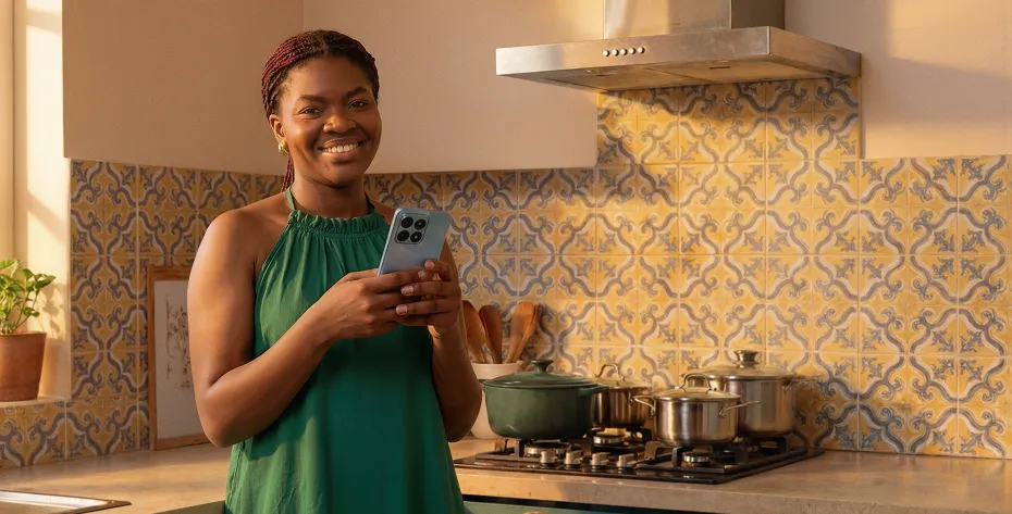 Smiling woman in green dress holding smartphone in kitchen with patterned tile backsplash and pots.