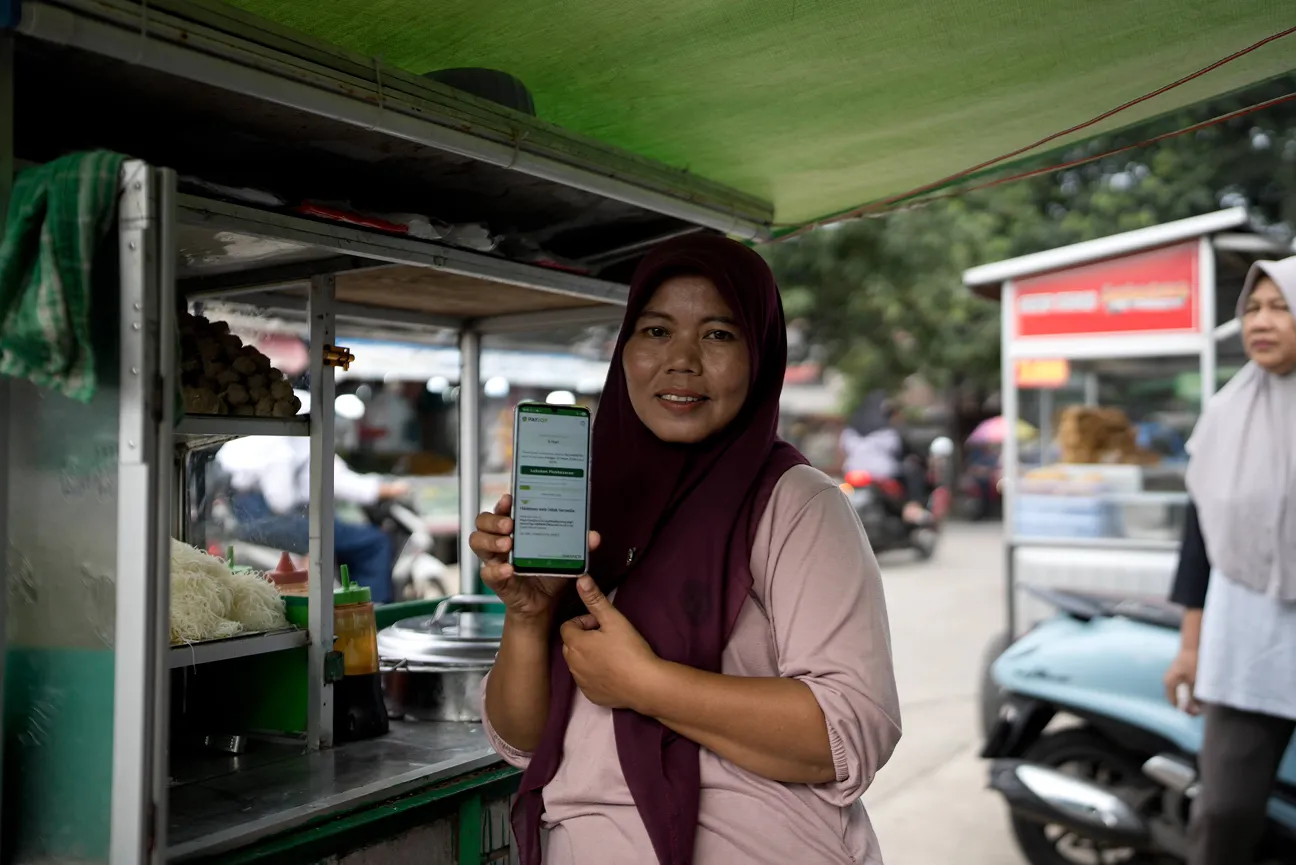 Woman in a maroon headscarf showing a smartphone with a payment app at a street food stall.