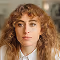 Portrait of a young woman with curly light brown hair and a calm expression against a neutral background.