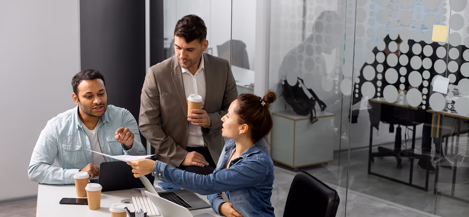 Three coworkers discussing a document at an office desk with laptops and coffee cups.