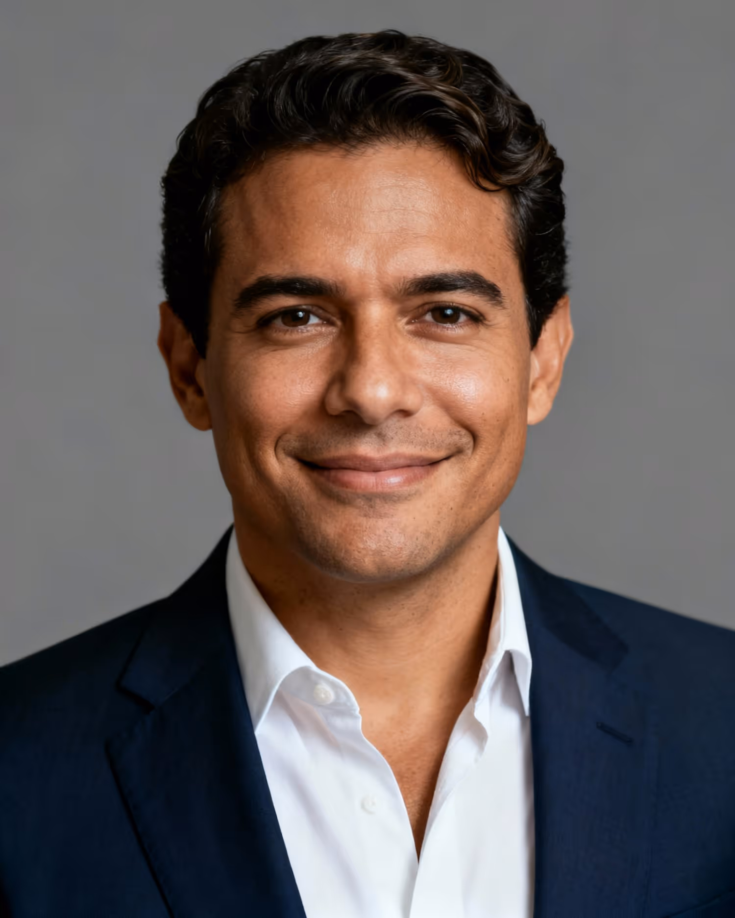 Smiling man with dark curly hair wearing a white shirt and dark suit jacket against a gray background.