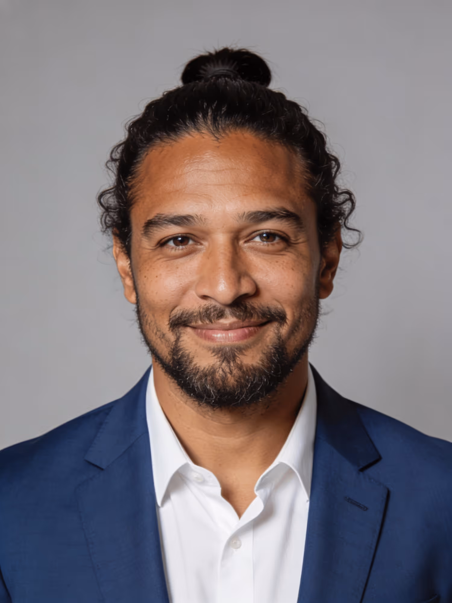 Smiling man with dark curly hair tied in a bun, wearing a blue suit jacket and white shirt against a gray background.