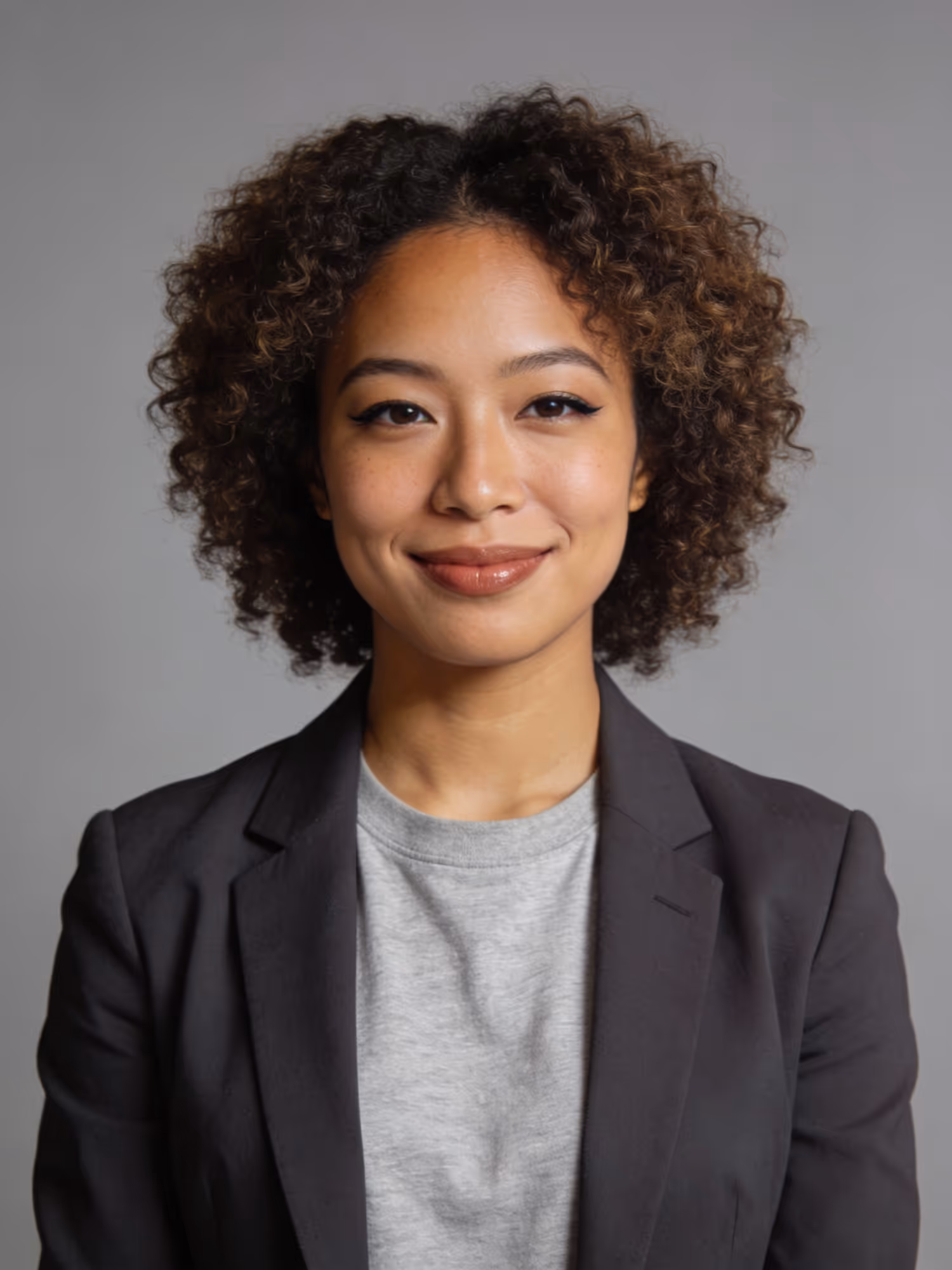 Smiling woman with curly hair wearing a gray shirt and black blazer against a gray background.