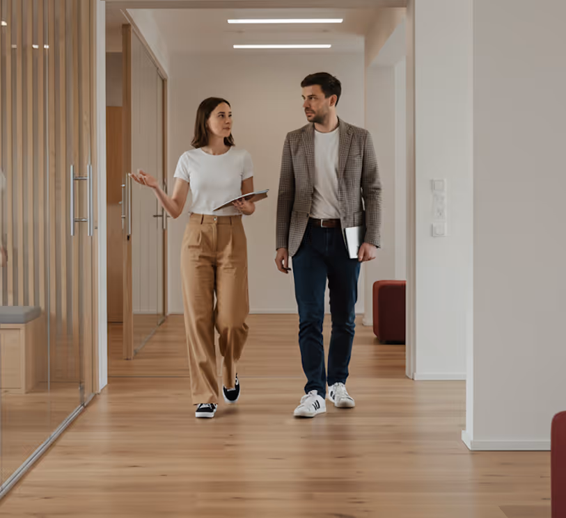 A woman and a man walking and talking together in a modern office hallway with wooden floors and glass walls.