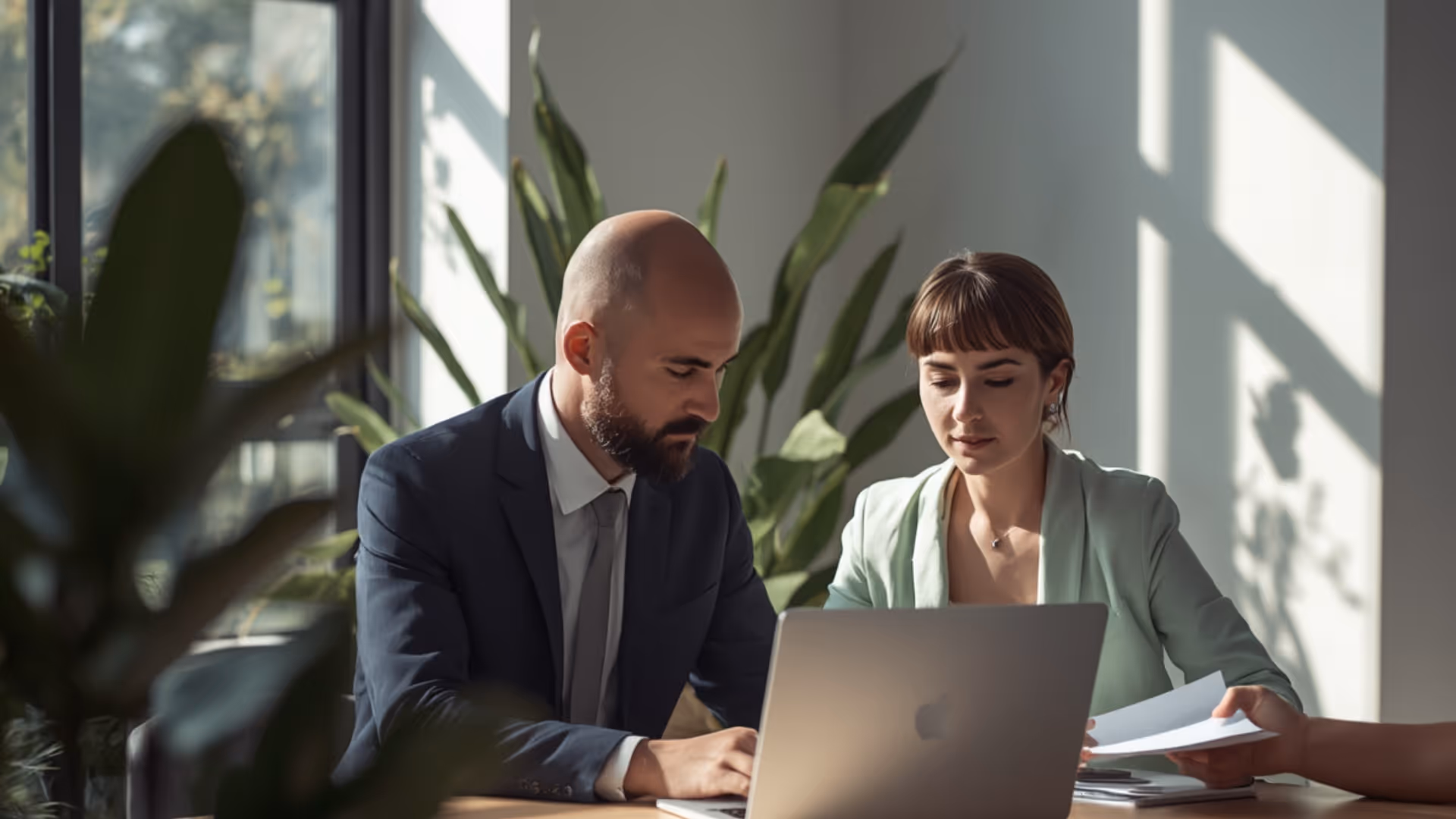 Two professionals working together at a desk with a laptop and documents in a sunlit office with plants.