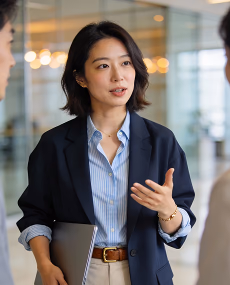 A professional woman with shoulder-length dark hair, wearing a navy blazer over a light blue striped shirt and beige pants, is speaking and gesturing with one hand while holding a laptop in her other hand in an office setting.