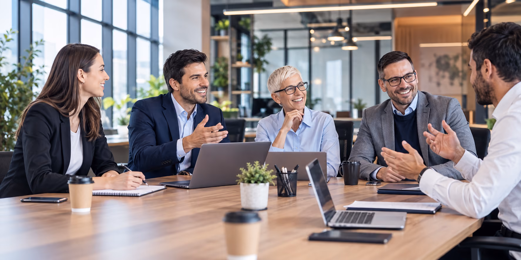 Five diverse business professionals sitting around a table in a modern office, engaged in a lively team meeting with laptops, notebooks, and coffee cups visible.