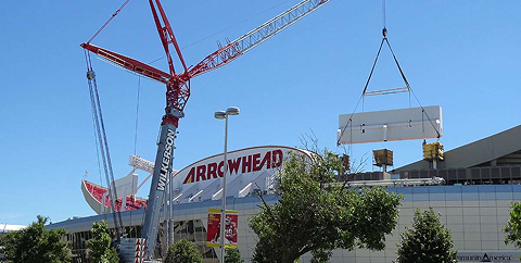 Construction crane lifting a large concrete slab near the Arrowhead Stadium under a clear blue sky.