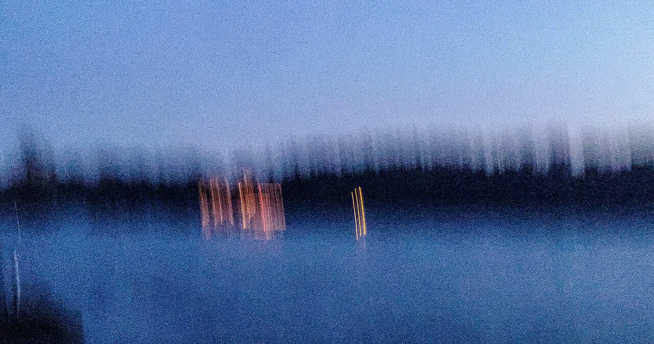 Blurry nighttime landscape with faint blue water and orange streetlights reflected in it, under a darkening sky.