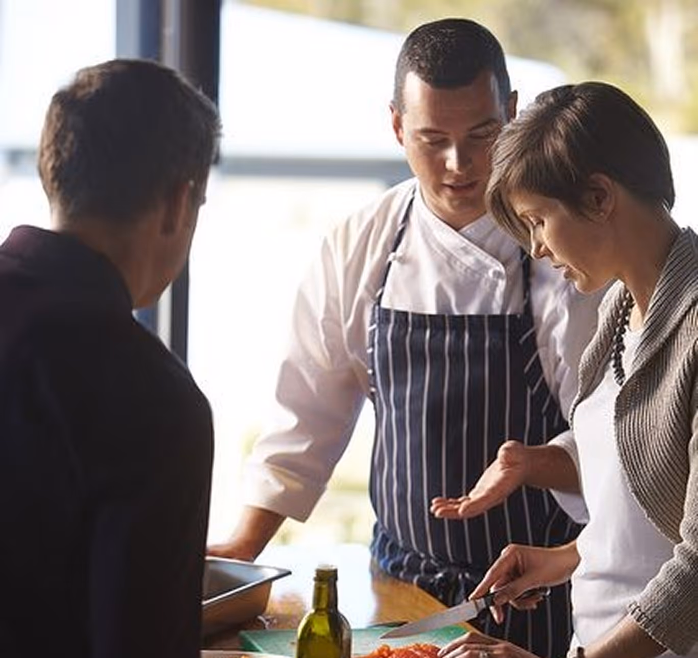 Un chef cuisinier en tablier rayé explique une technique à une femme qui coupe des légumes sur une planche à découper devant un homme observant.