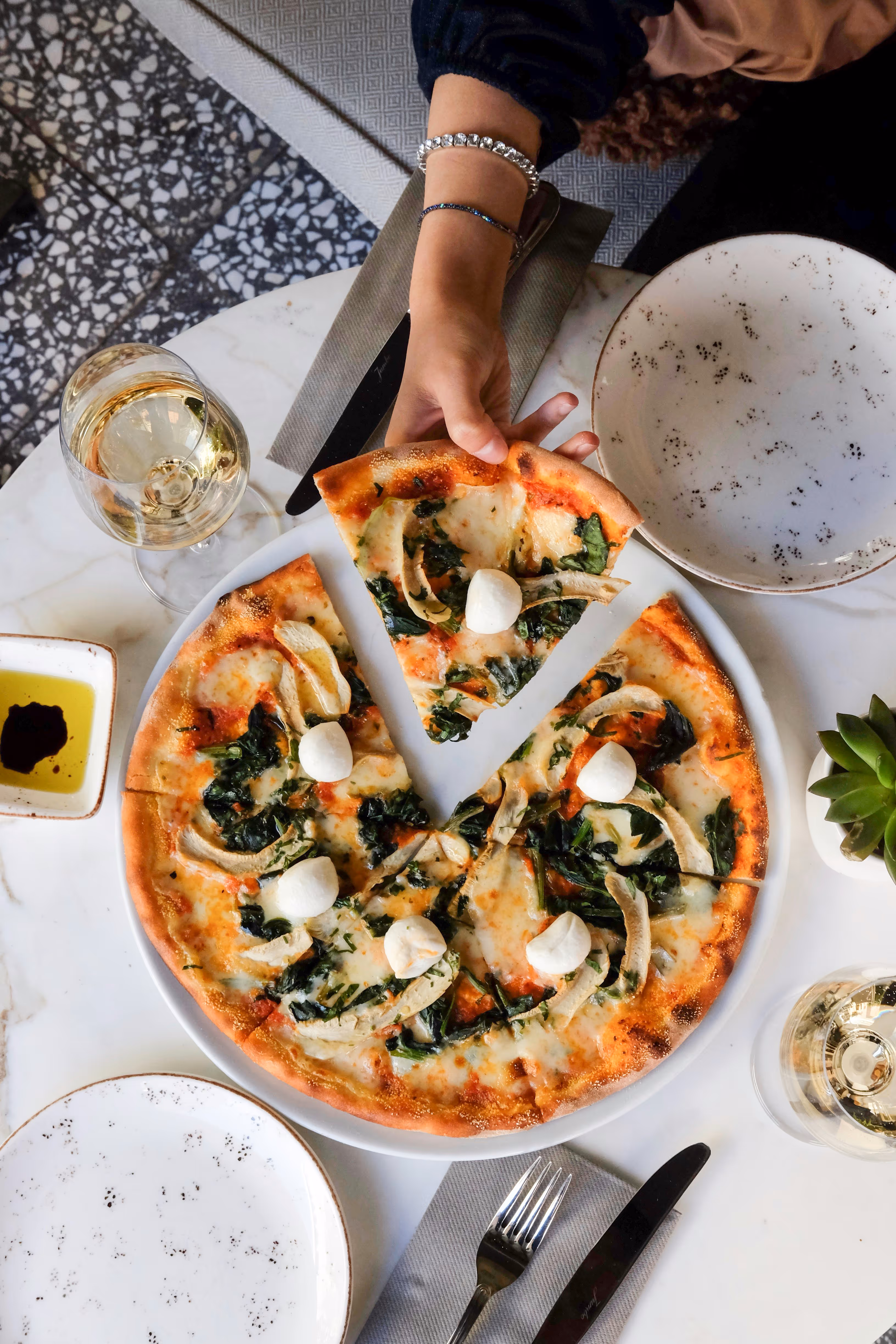 Une main saisit une part de pizza aux légumes verts et boules de mozzarella sur une table avec deux verres de vin blanc et assiettes vides.