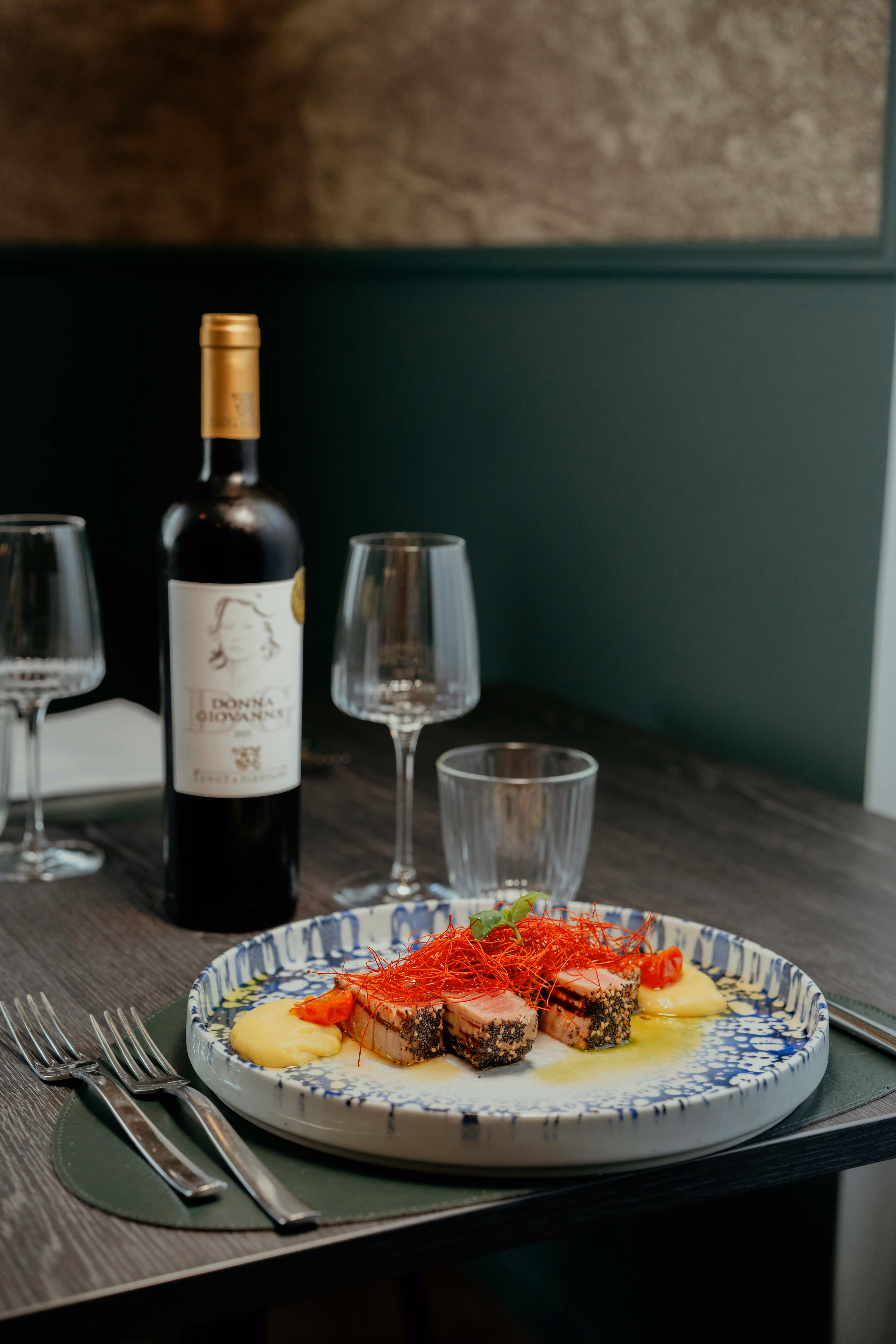 Plat gastronomique avec médaillons de viande garnis de fils rouges sur une assiette décorée, accompagnés de purée et de morceaux de tomate, avec une bouteille de vin rouge et deux verres vides en arrière-plan.