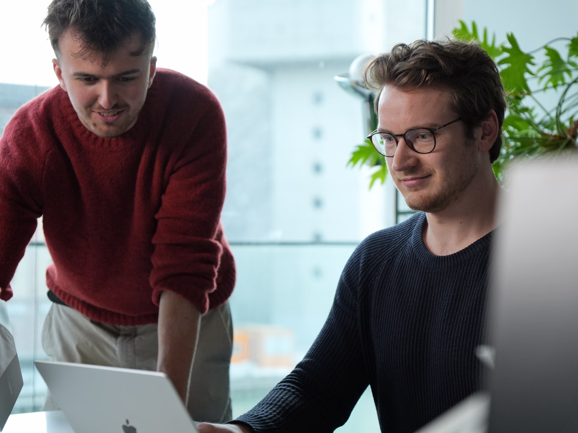Two men working together at a laptop in a modern office with large windows and plants.
