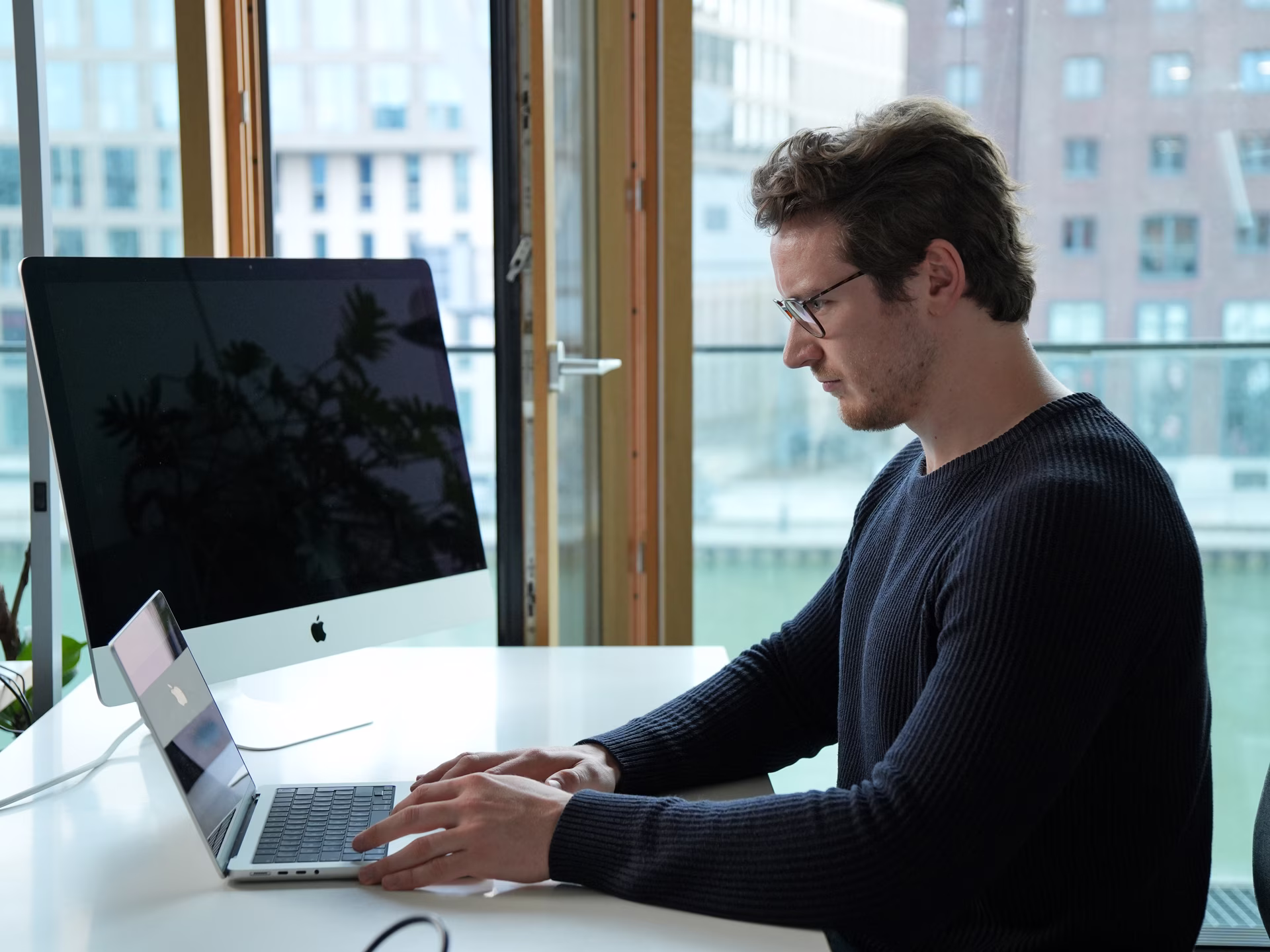 Man in glasses typing on a laptop at a desk with a large monitor and window in the background.
