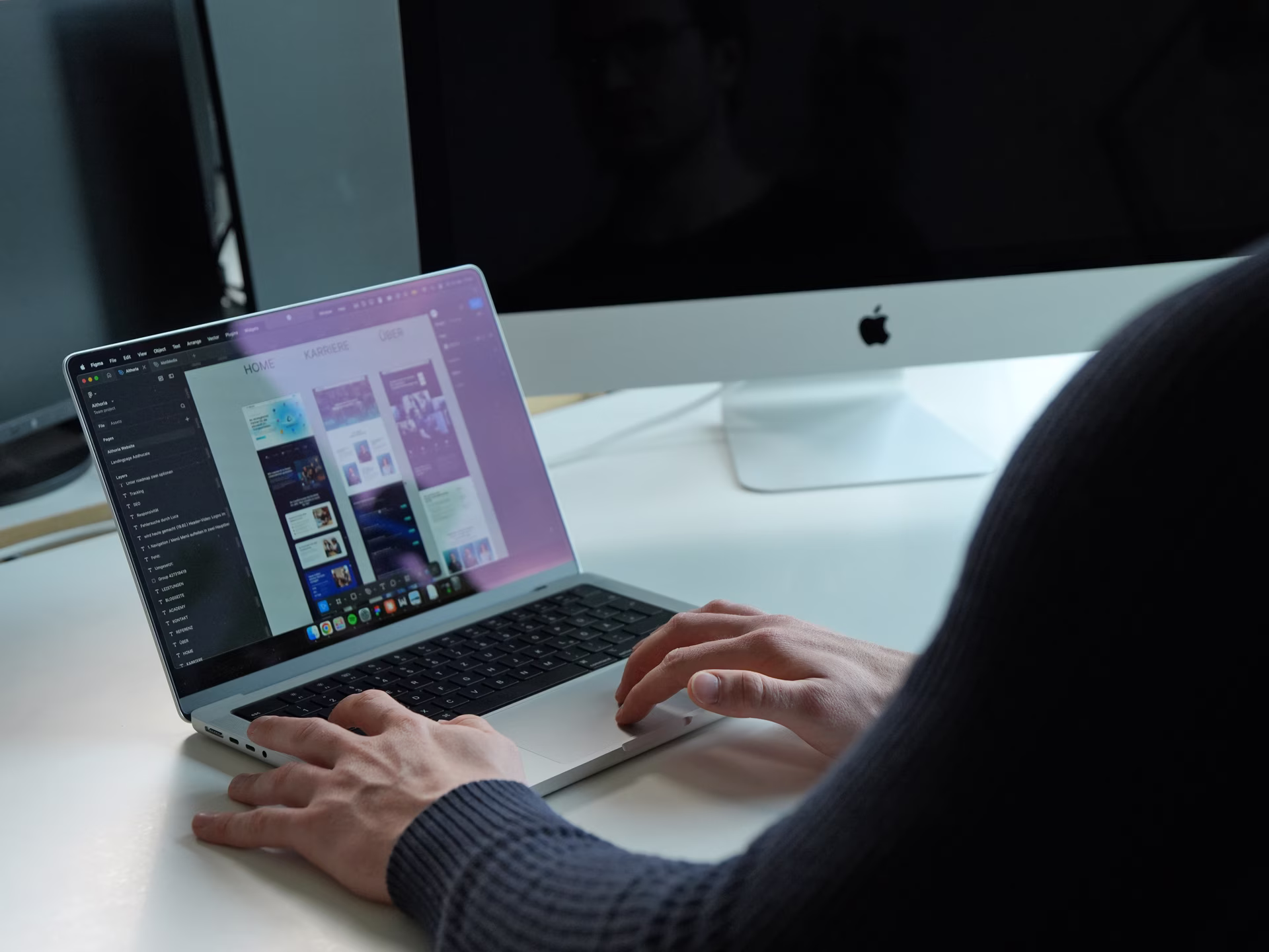 Person working on a laptop displaying a design project, with an iMac in the background.