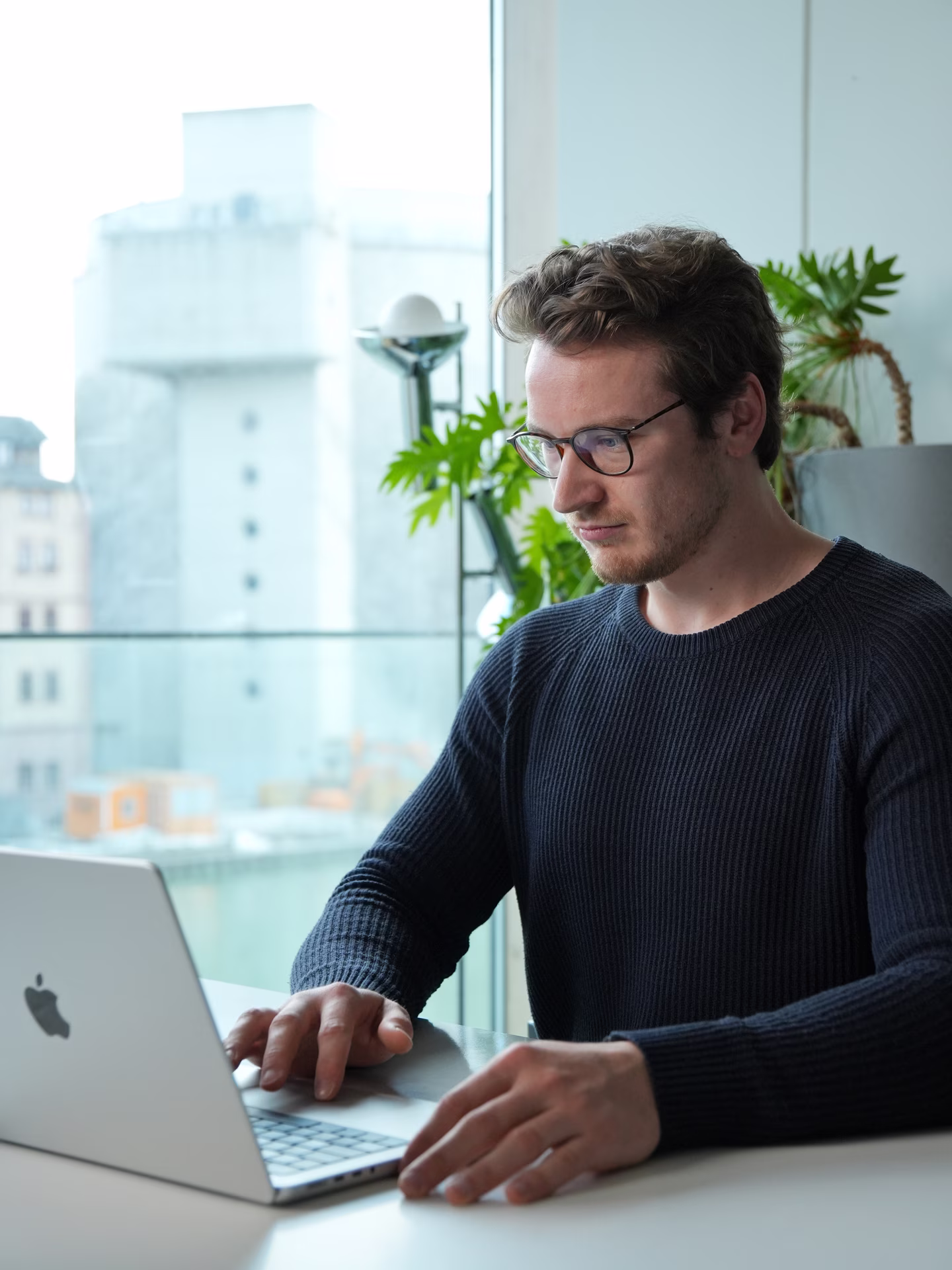 Man wearing glasses and a dark sweater working on a laptop at a desk by a window.