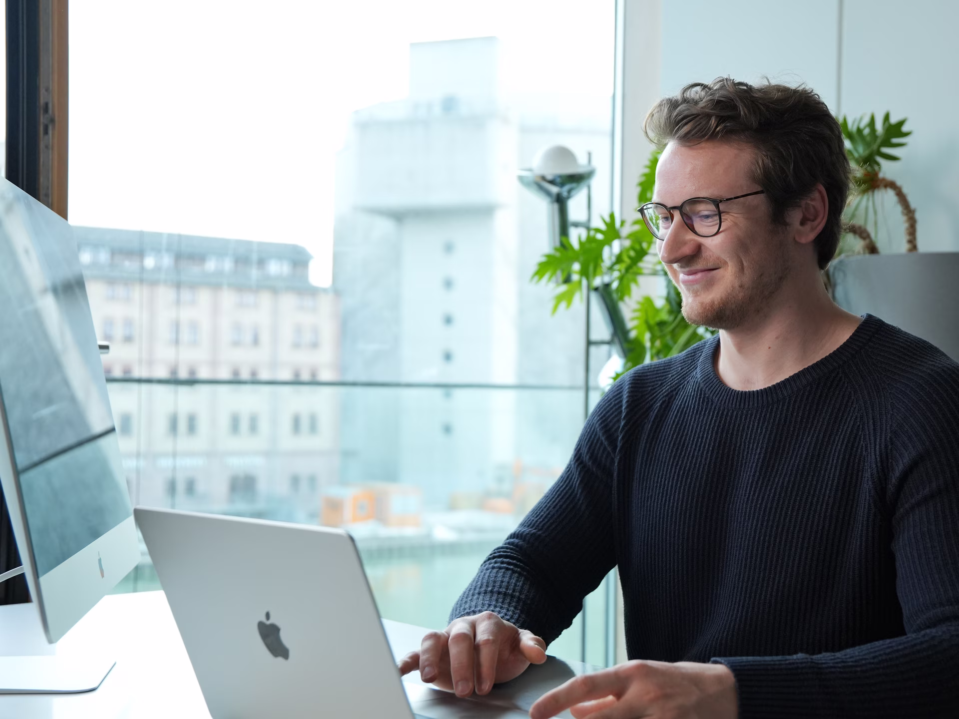 Smiling man wearing glasses working on a laptop at a desk with a large window and plants behind him.