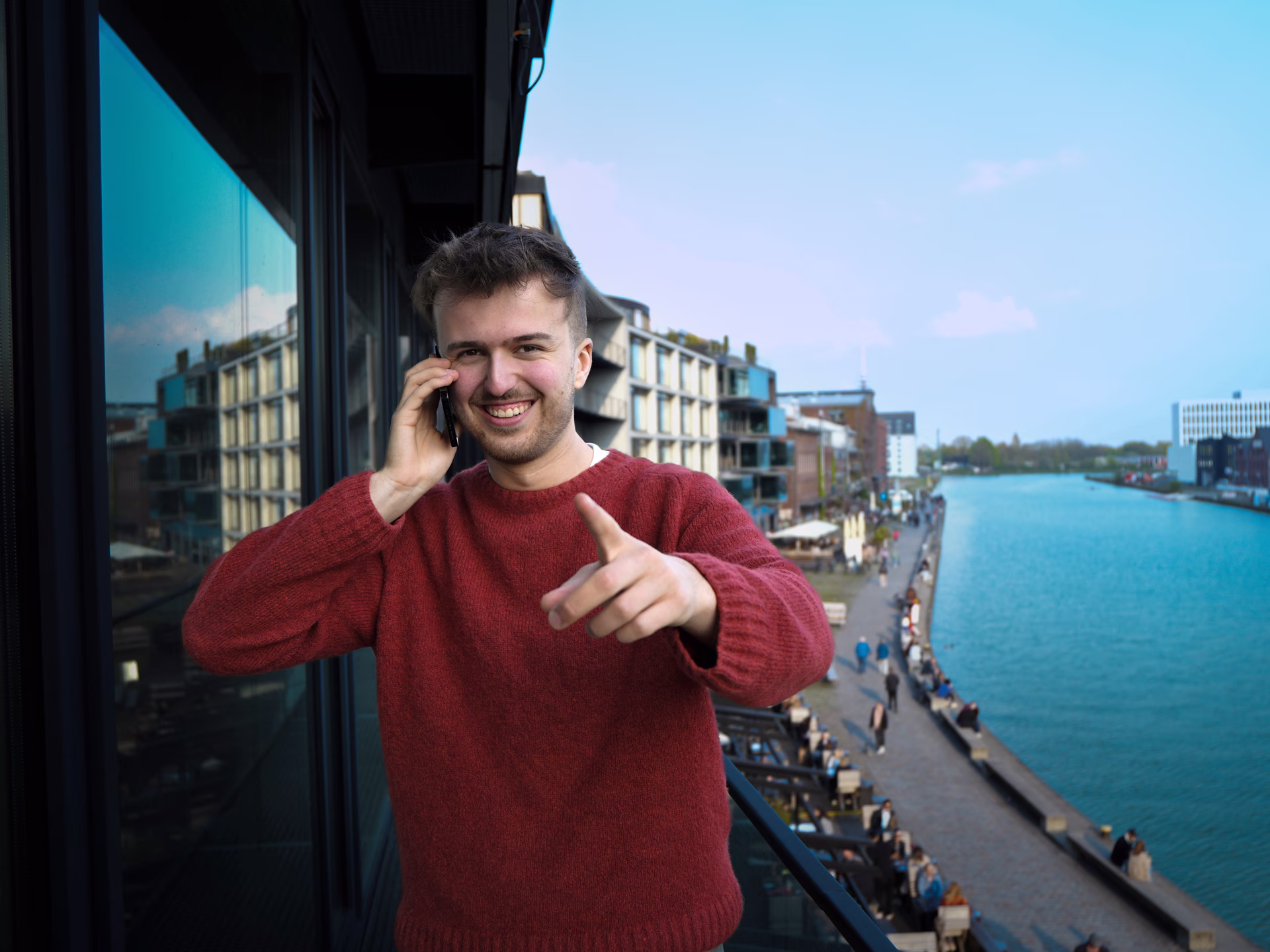 Smiling man in red sweater talks on phone and points forward on a balcony by a riverwalk.