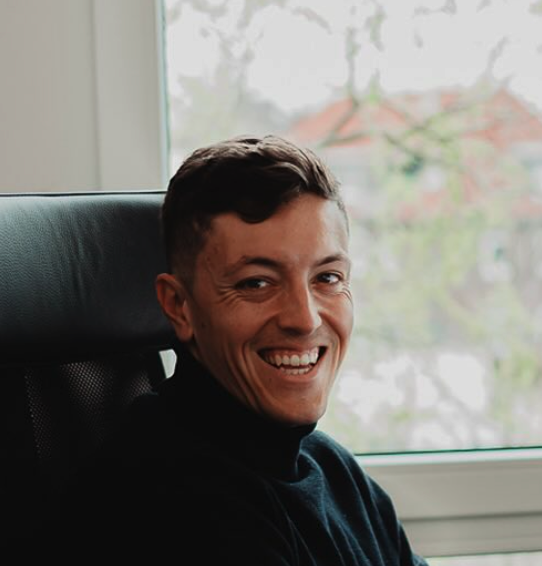 Smiling man wearing a black turtleneck sitting in a black office chair by a window.