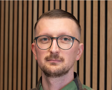 Man with glasses, light beard, and short hair, wearing a green shirt with striped wood background.