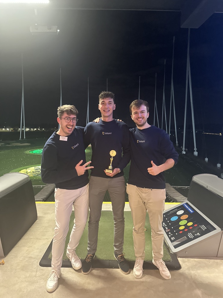 Drei junge Männer in Matching-Pullovern halten und zeigen stolz eine Trophäe nachts an einem Golfplatz.