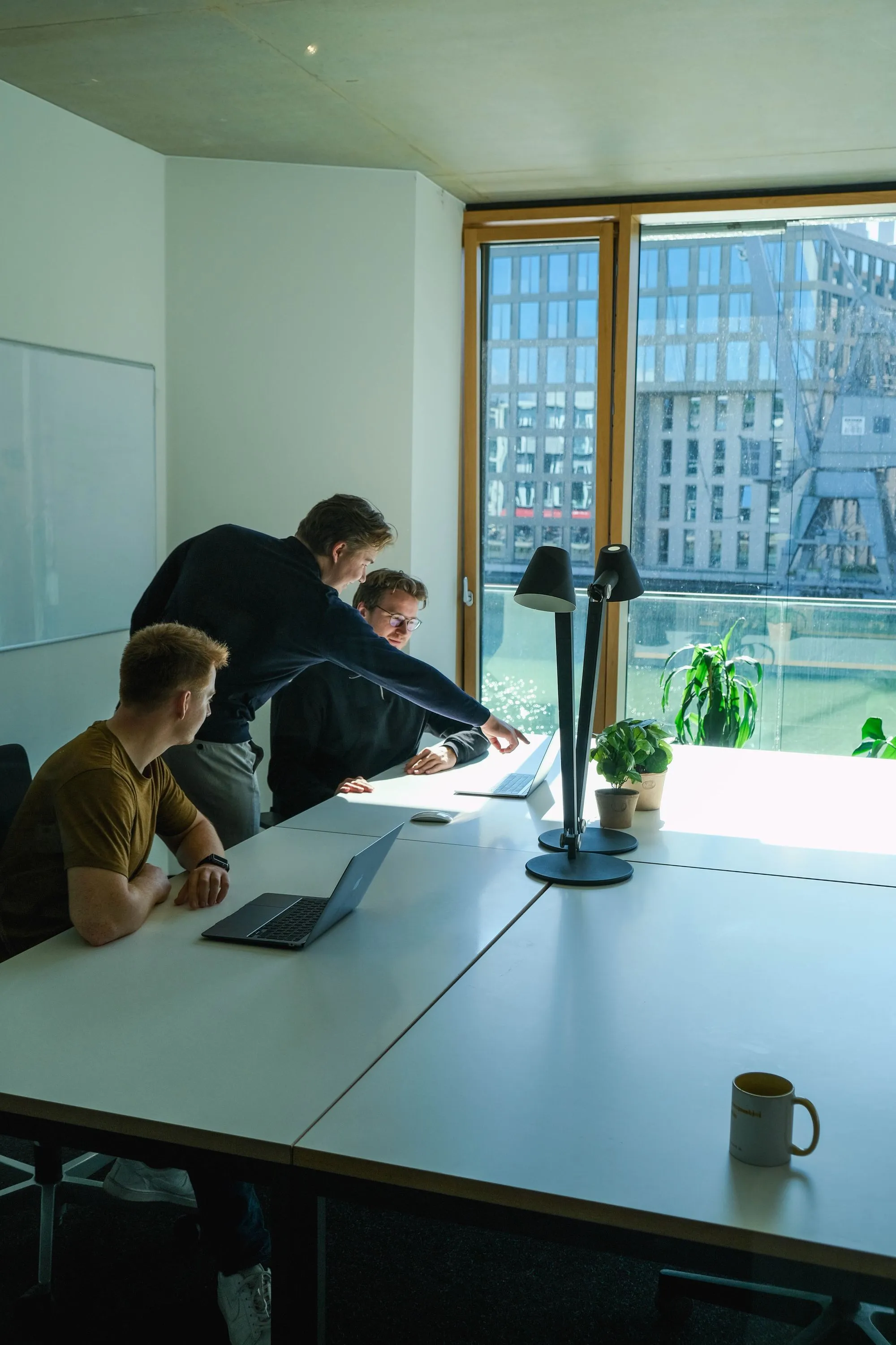 Drei Männer arbeiten an Laptops an einem Tisch vor großen Fenstern mit Blick auf ein modernes Gebäude.