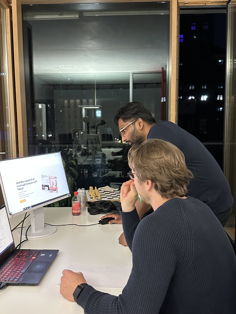 Zwei Männer sitzen und stehen nachts am Schreibtisch und schauen auf Monitore im Büro.