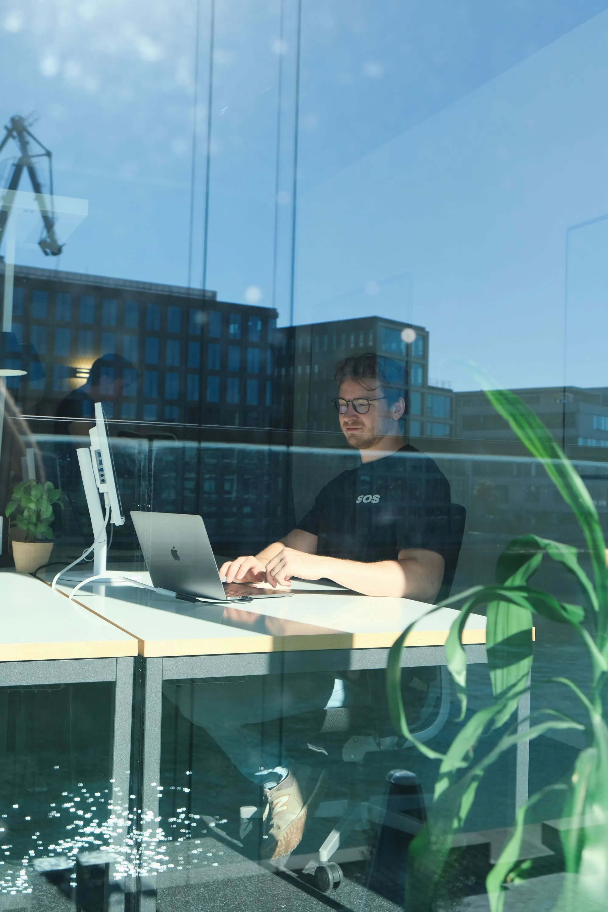 Mann mit Brille arbeitet an einem Laptop an einem hellen Schreibtisch in einem Büro mit Fensterblick.