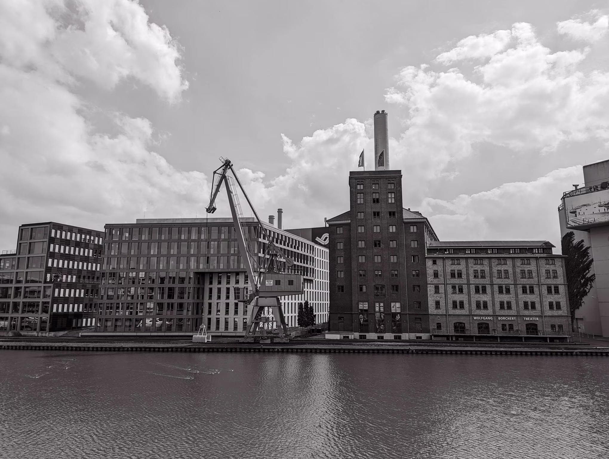 Schwarz-weiß-Bild eines Hafenbeckens mit Industriebauten, Kran und Theatergebäude am Wasser.