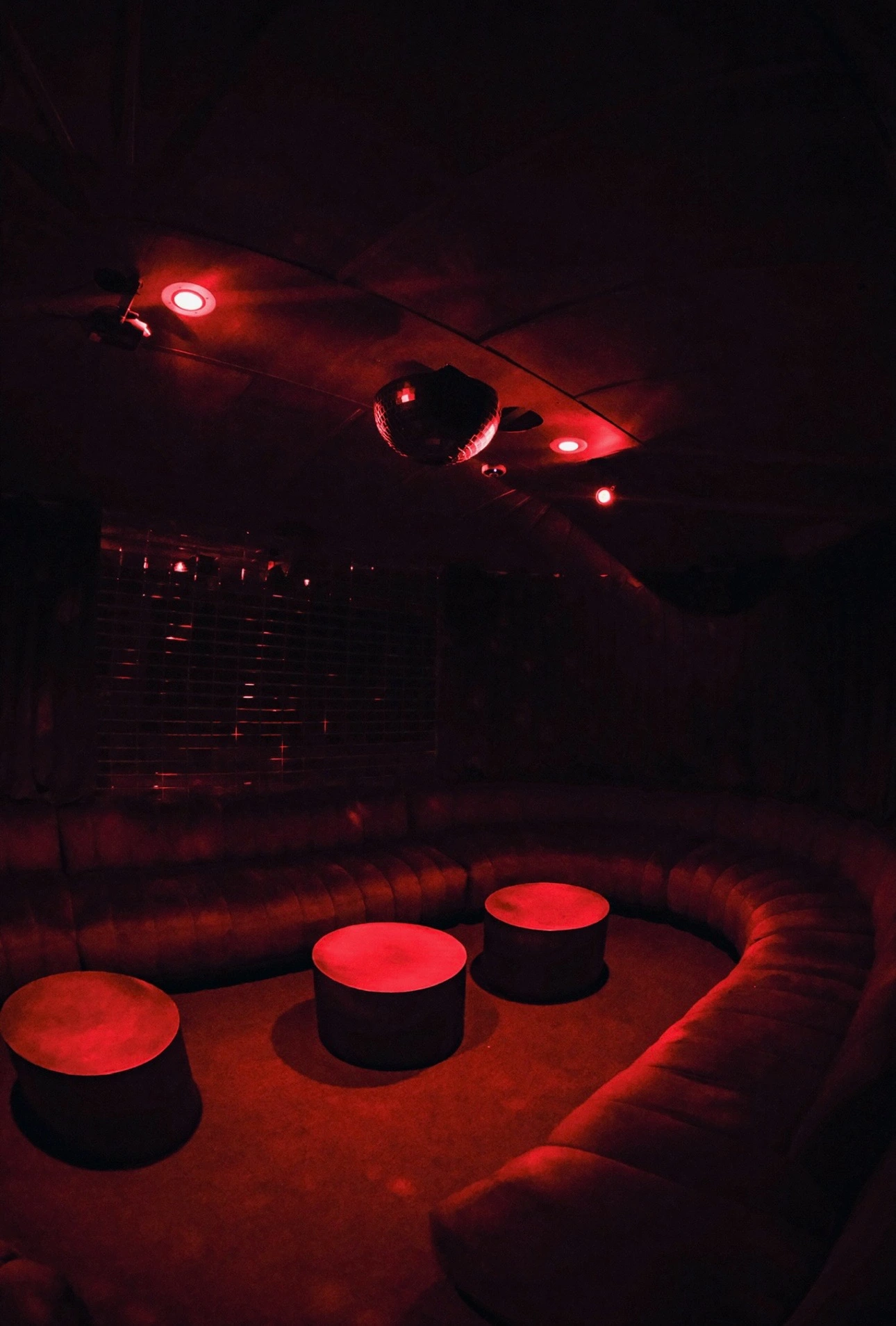 Dimly lit lounge with curved sofa, round tables, and red ambient lighting under a disco ball.
