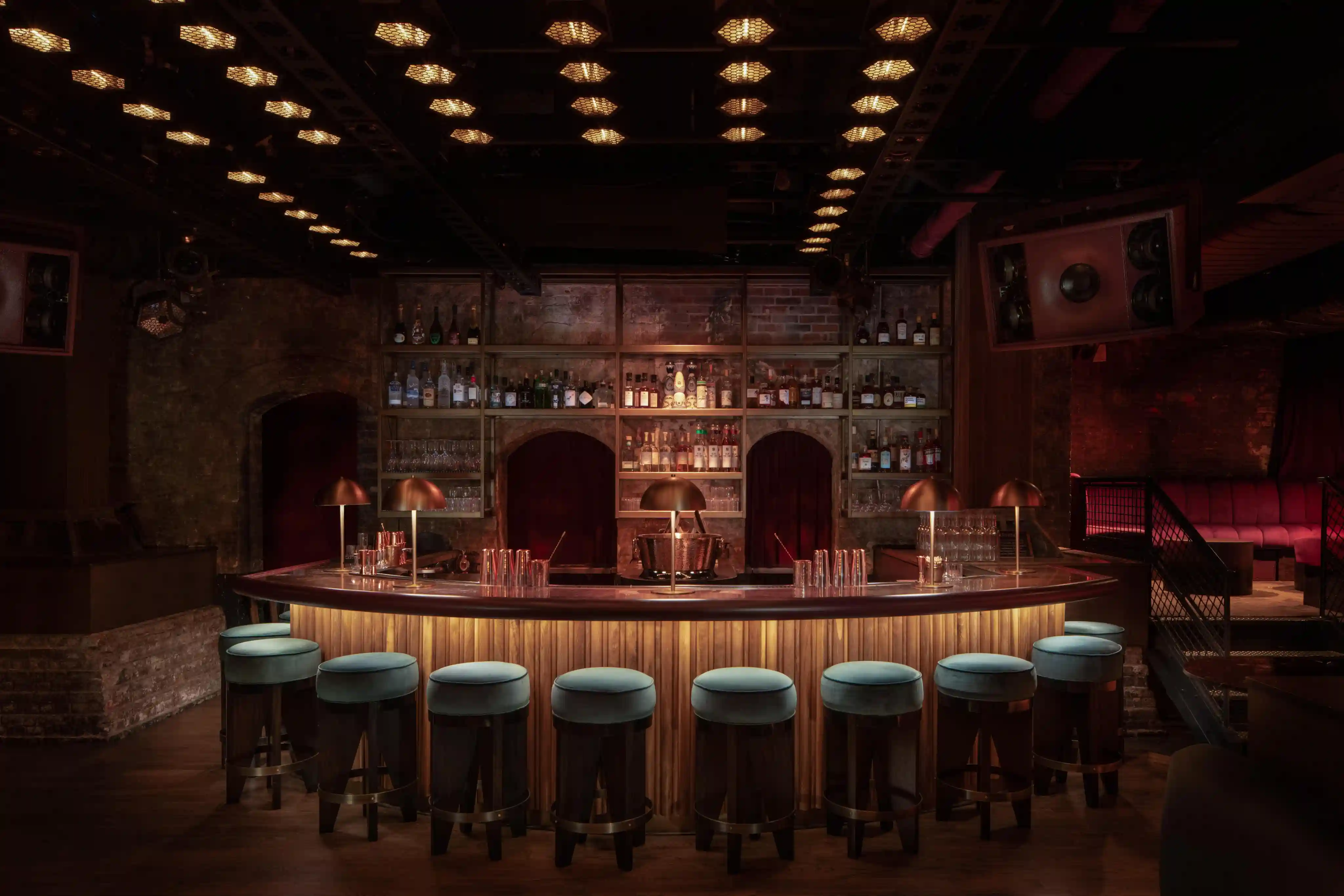 Dimly lit bar with a curved counter, blue cushioned stools, and shelves of liquor bottles on a brick wall.