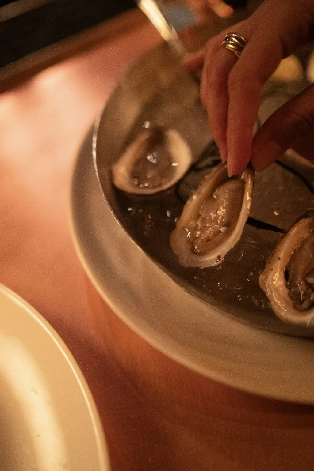 Hand with rings picking a fresh oyster from a plate on a wooden table.