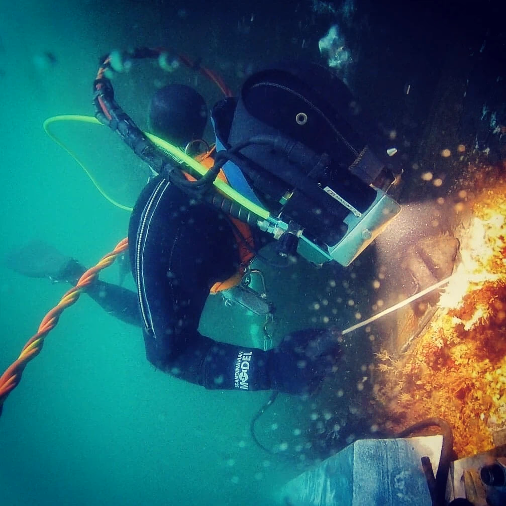 Dykker som arbeider under vann med utstyr og kabel nær en rusten struktur eller vrak.