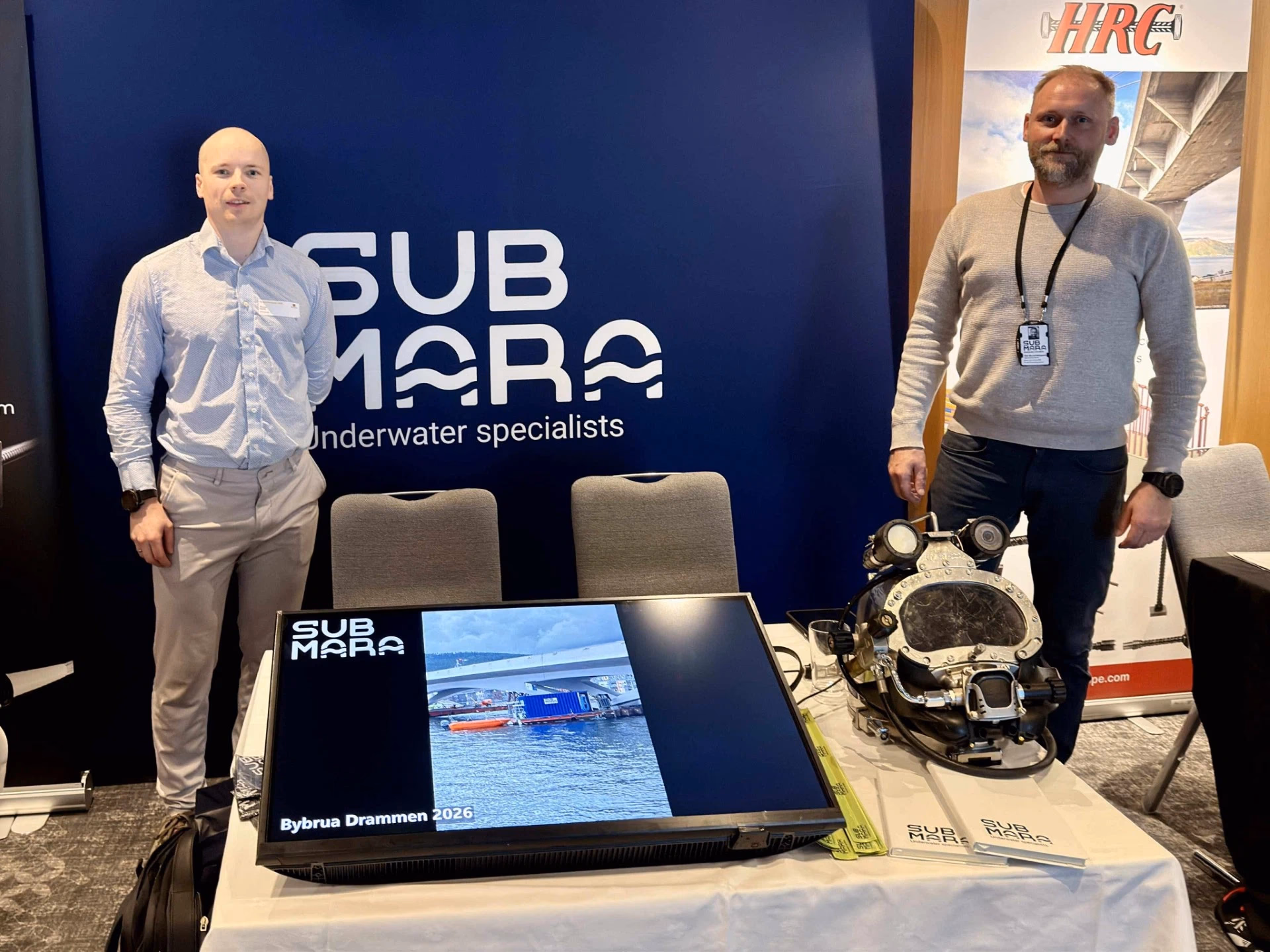 To menn står ved en stand for SubMara med dykkermaskin og en skjerm som viser en bru.