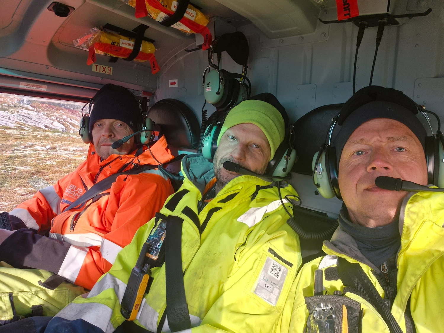 Tre menn i vernejakker og headset sitter side om side i helikopterets cockpit.