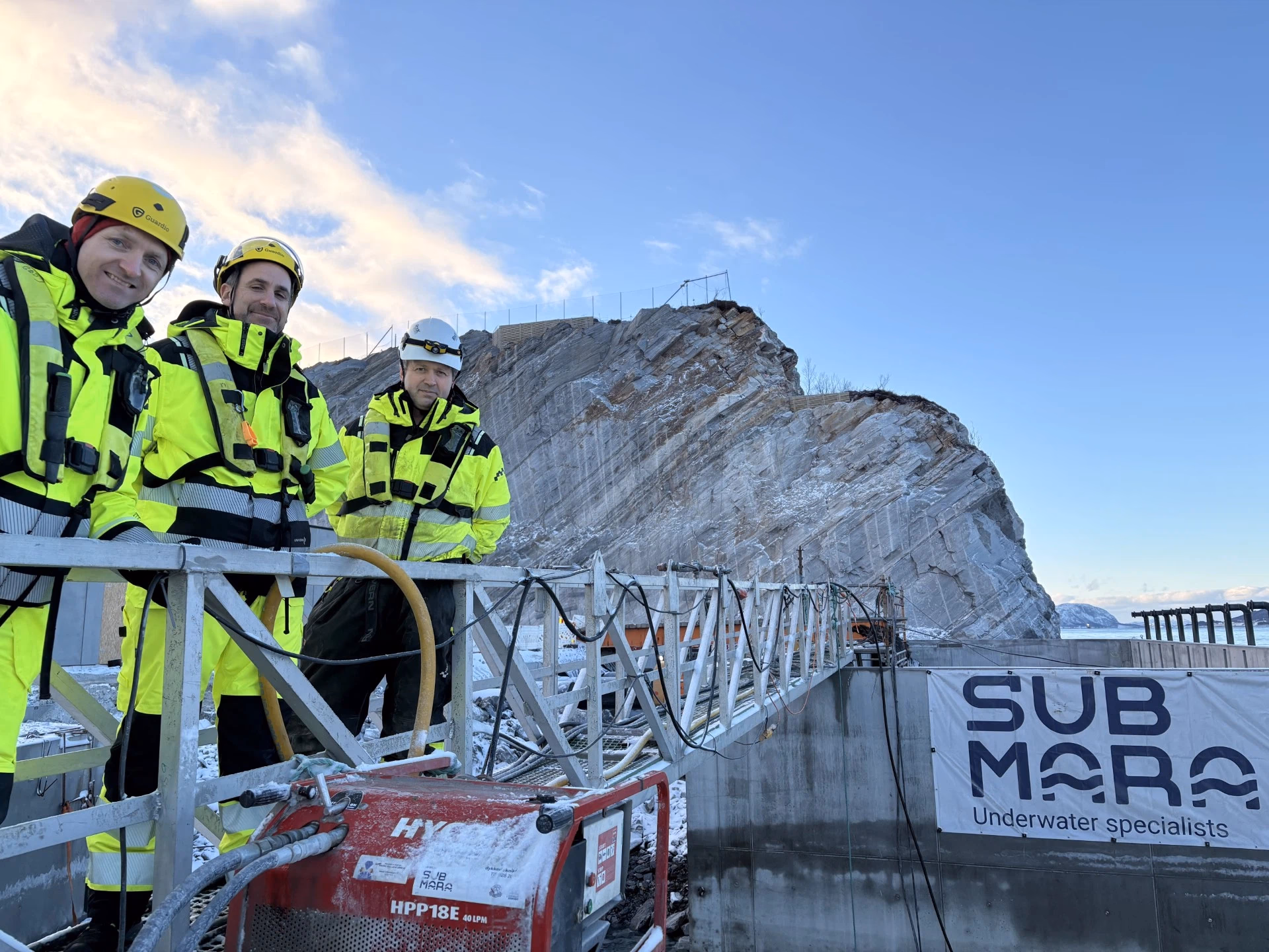 Tre arbeidere i gule vernedrakter og hjelmer ved en arbeidsplattform foran en fjellformasjon og skilt med SubMara-logo.