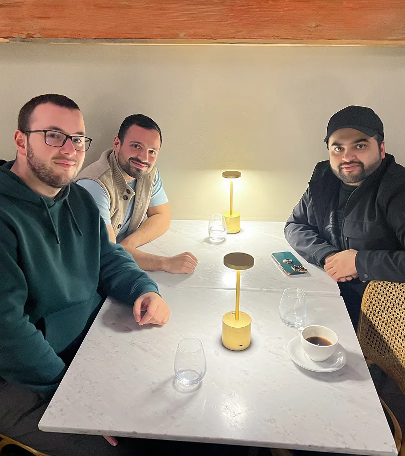 Three men sitting around a white marble table with two small modern lamps, glasses, a coffee cup, and a smartphone.