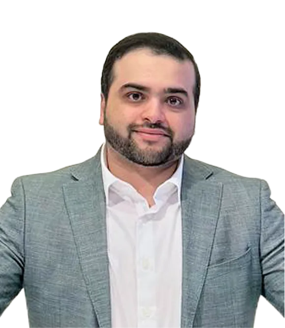 Man with short dark hair and beard wearing a light gray suit jacket over a white dress shirt, smiling slightly.
