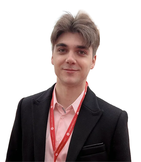 Young man with light brown hair wearing a black blazer, pink shirt, and red lanyard.