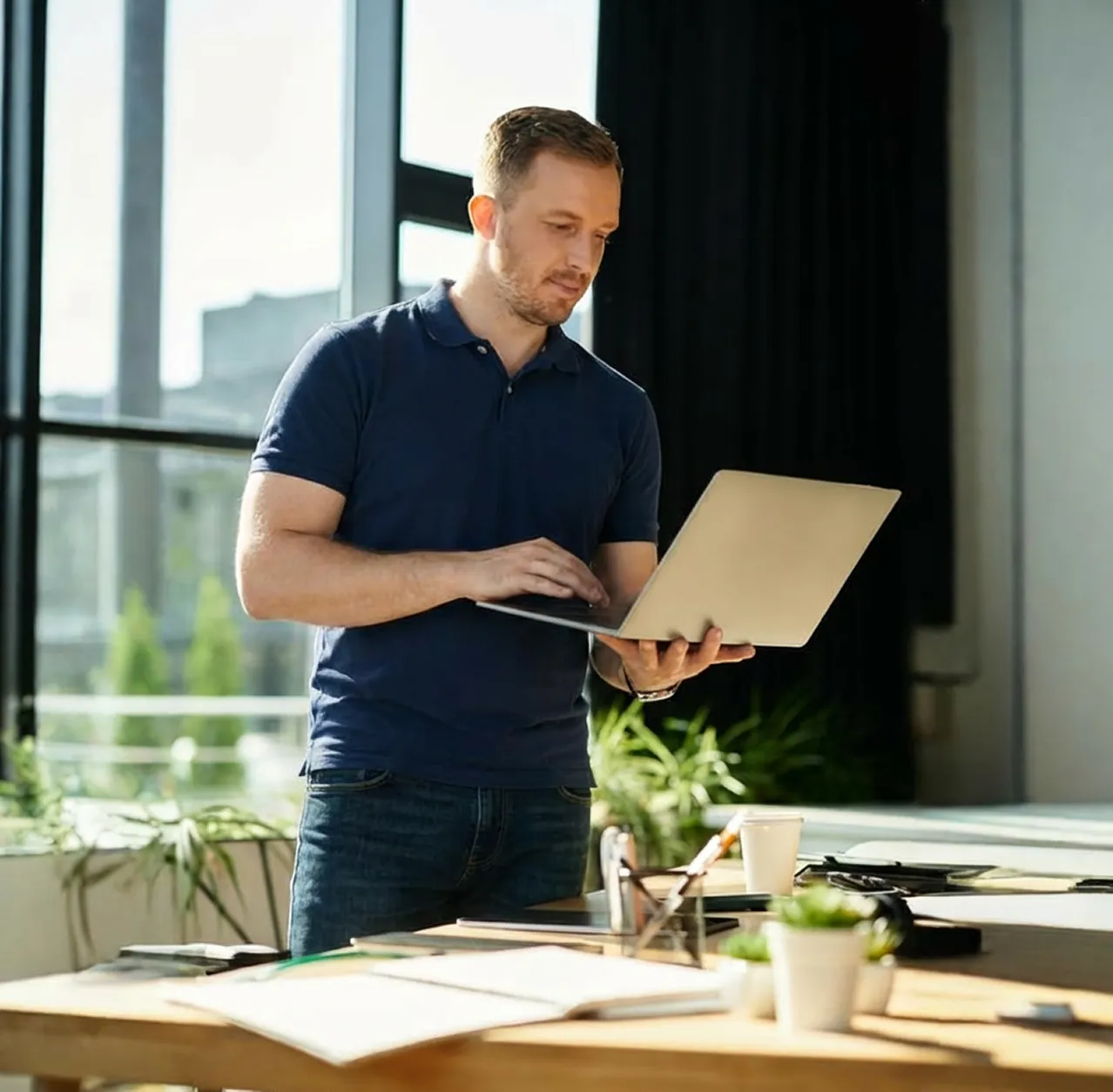 Man in a navy blue polo shirt working on a laptop in a bright office with plants and papers on the desk.