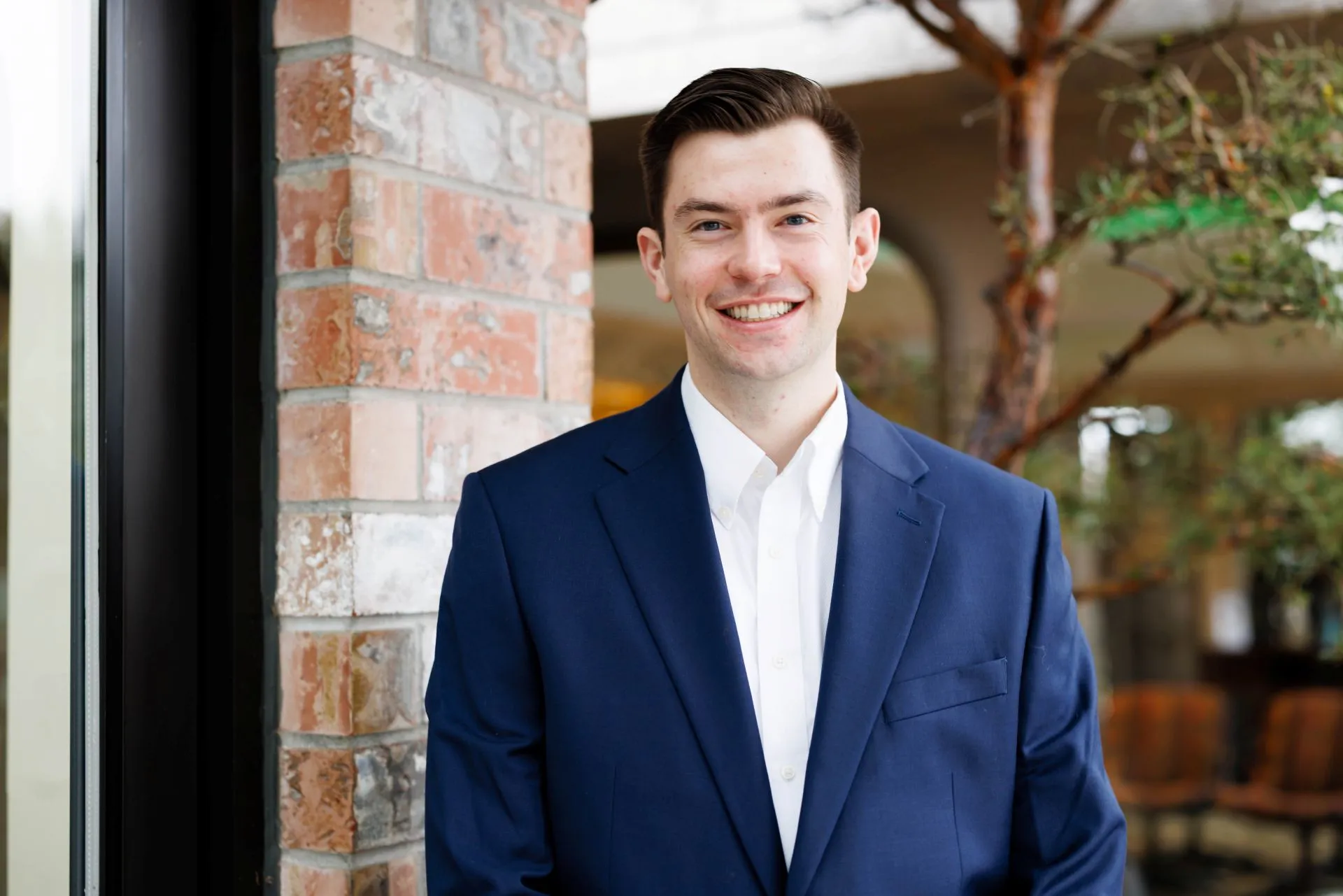 Smiling man in a blue suit jacket and white shirt standing indoors near a brick wall.