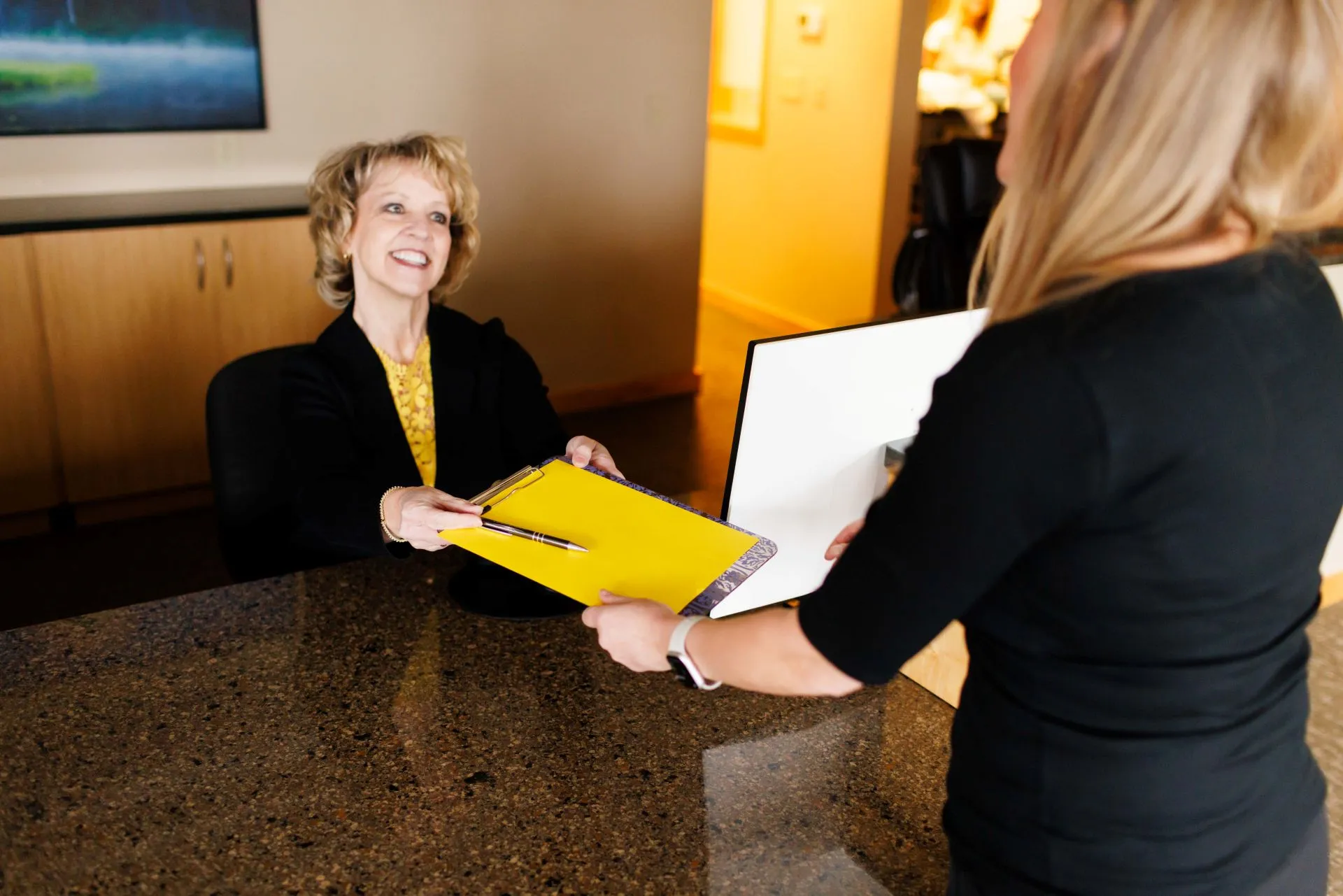 Receptionist handing a clipboard with a yellow paper and pen to a visitor at a front desk.