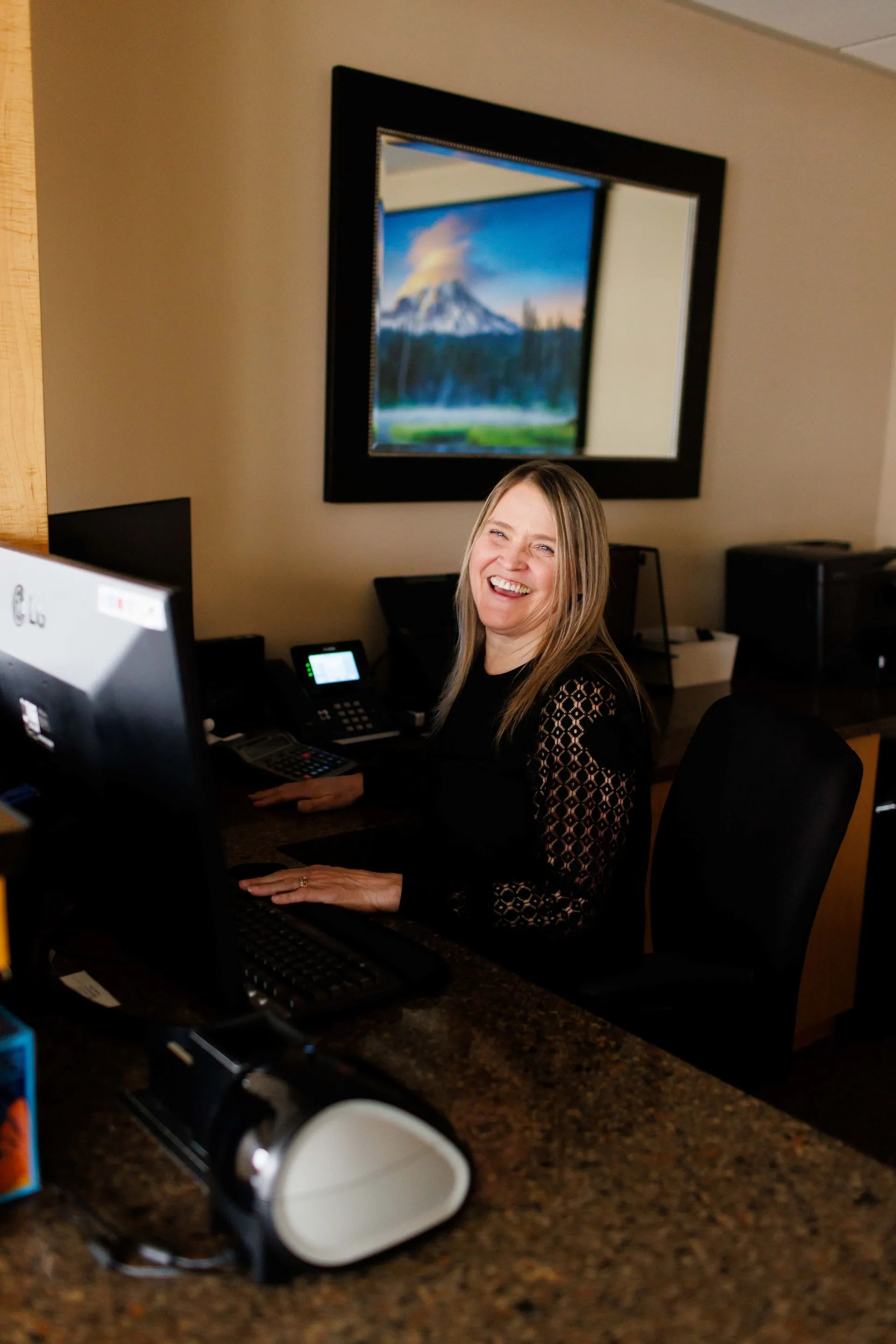 Smiling woman with long blonde hair sitting at a desk in front of a computer in an office.