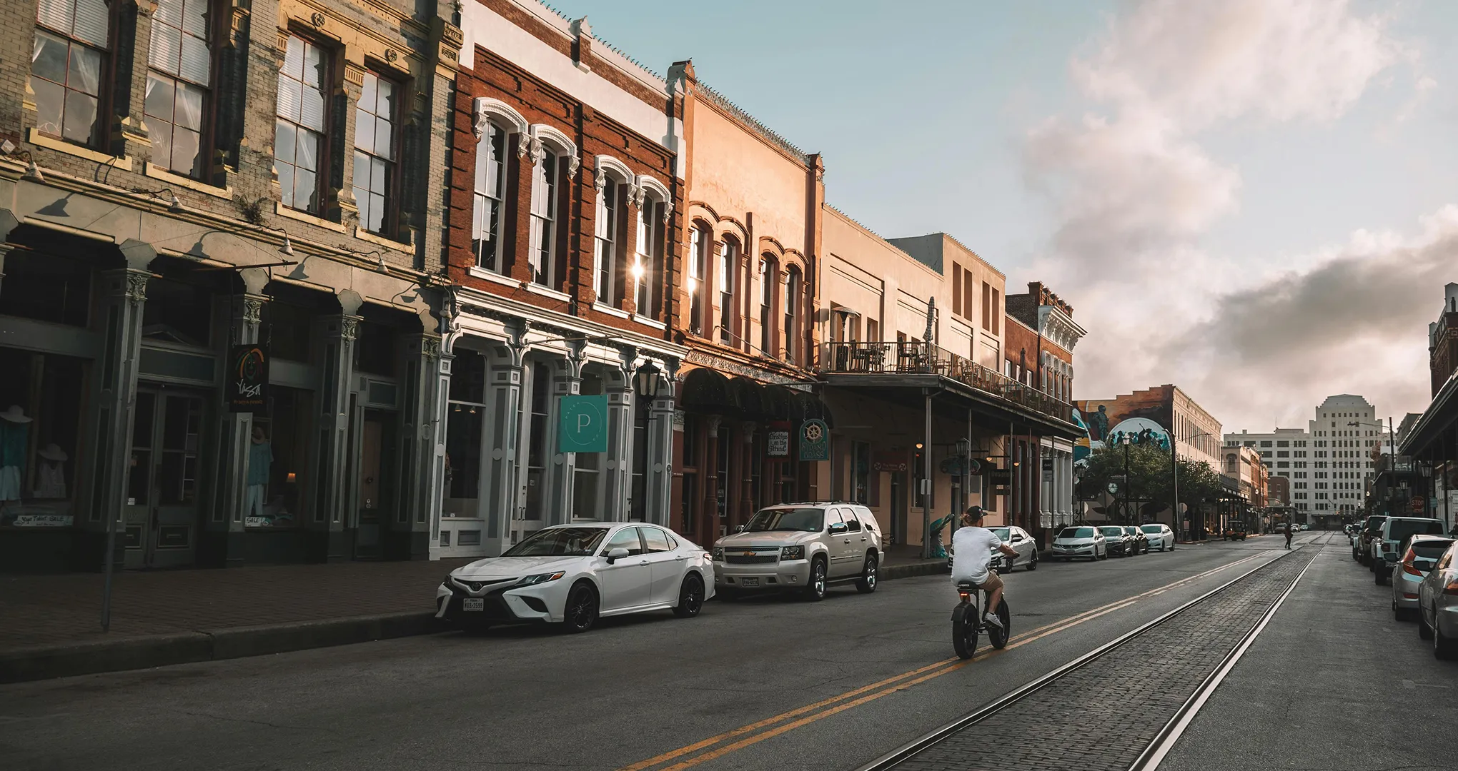 Urban street scene with historic buildings, parked cars, and a person riding a bicycle on rails.