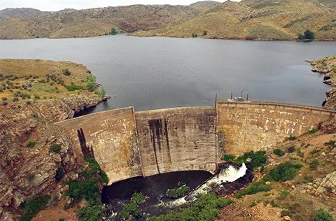 Aerial view of a concrete dam holding back a large reservoir with surrounding rocky hills.