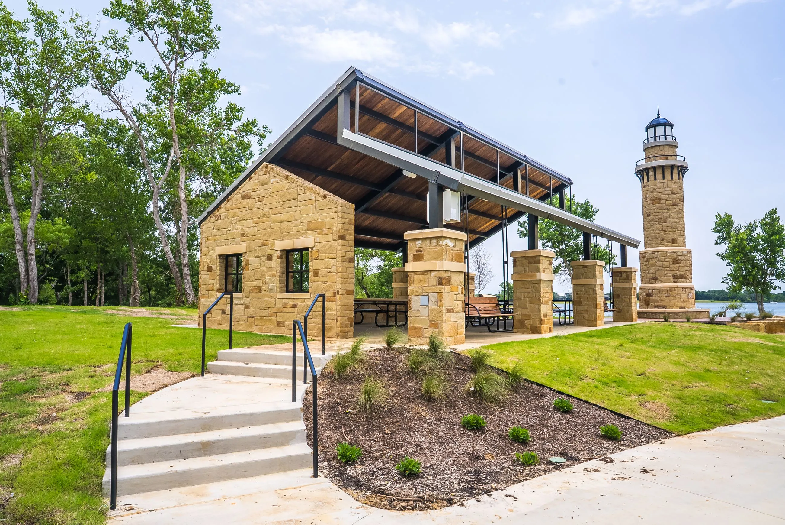 Stone pavilion with picnic tables and benches near a lakeside lighthouse in a green park area.