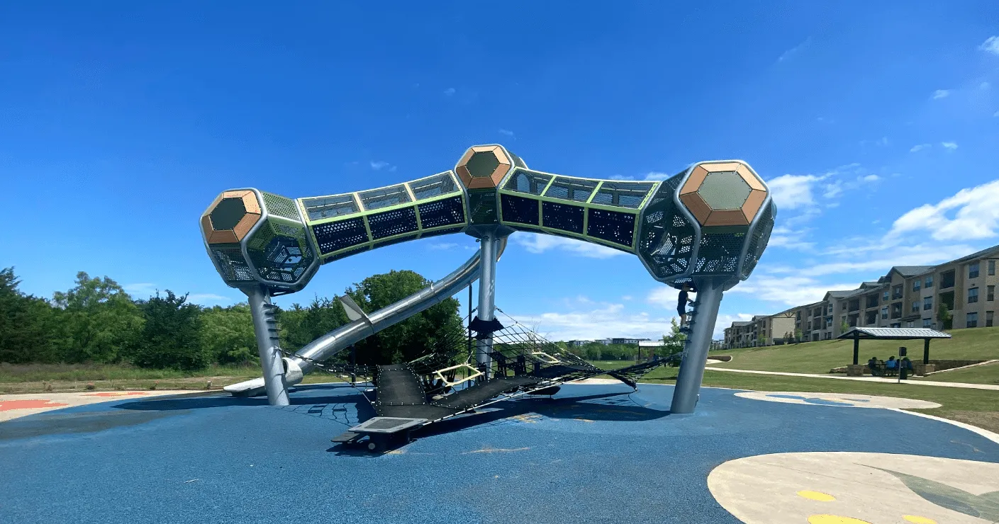 Modern playground structure with interconnected hexagonal pods, a slide, and climbing nets on a blue rubber surface under a clear sky.