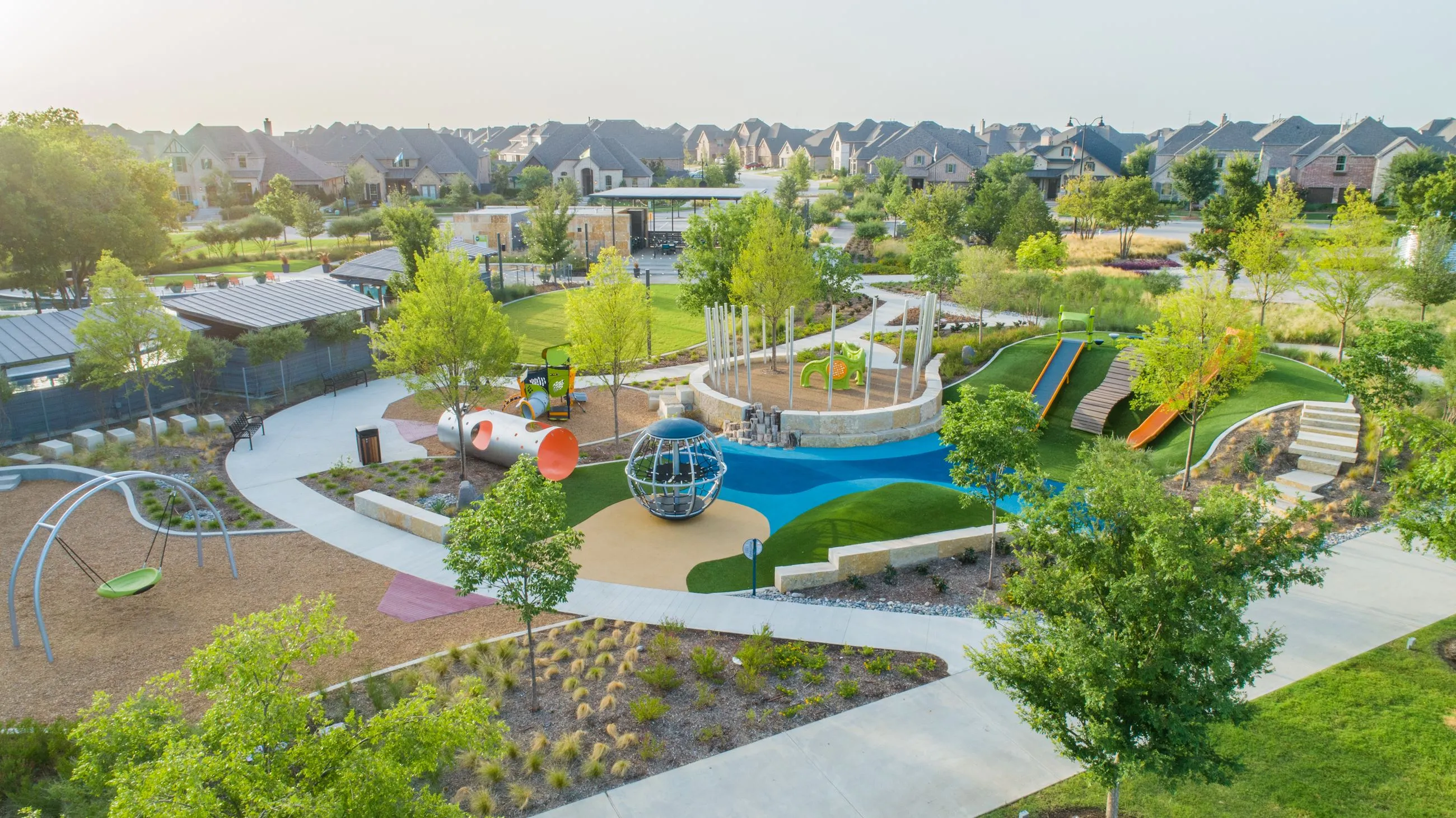 Aerial view of a colorful playground with slides, climbing structures, swings, and walking paths surrounded by green trees and residential houses.