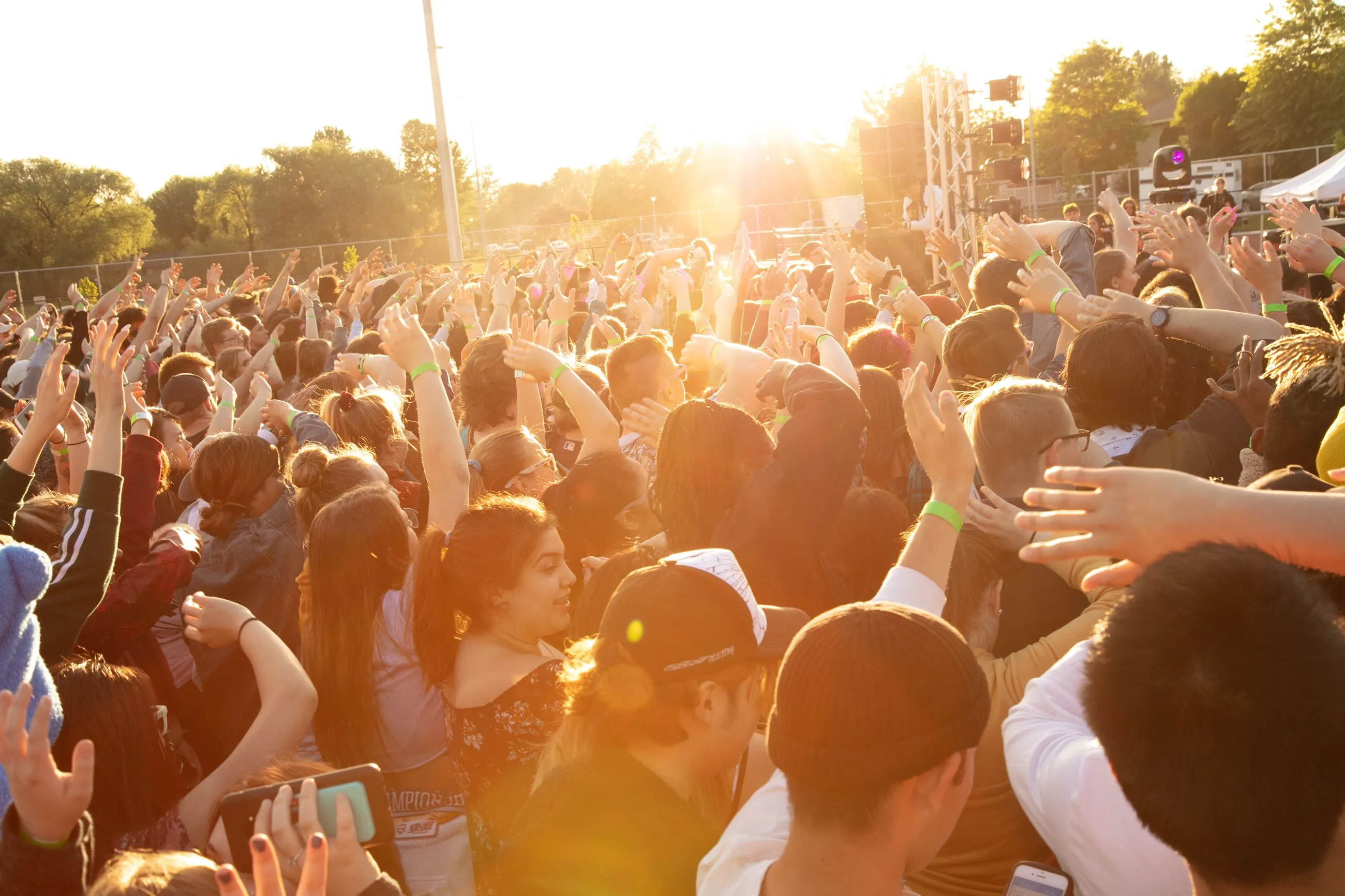 Crowd of people at an outdoor event raising their hands, illuminated by warm sunlight.