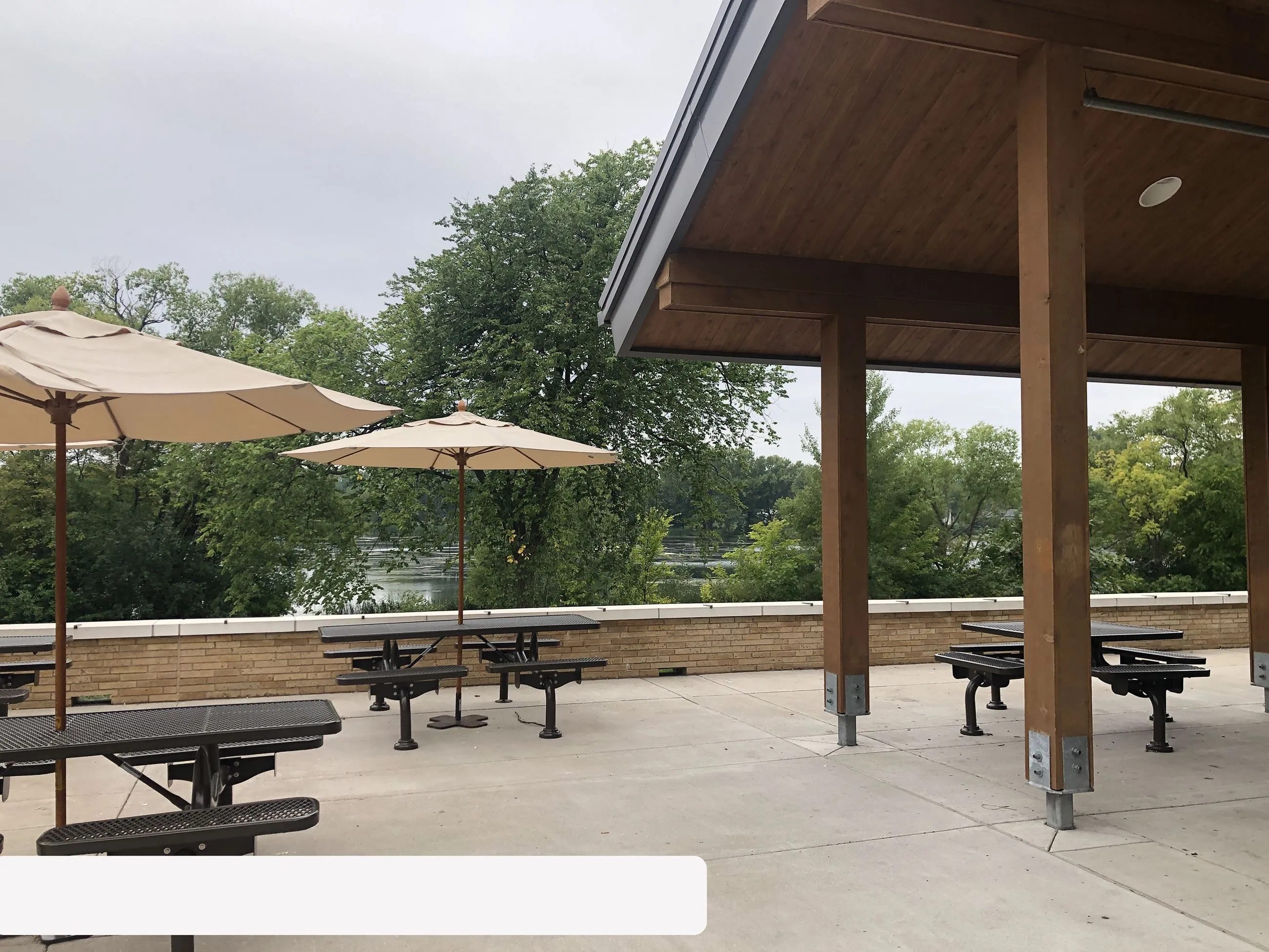Outdoor park seating area with picnic tables, umbrellas, a wooden shelter, green trees, and a river in the background.